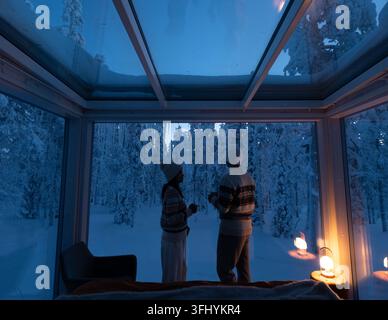 Due persone si godono un momento di pace all'interno di un'accogliente cabina di vetro, osservando il tranquillo paesaggio innevato di Akaslompolo, Lapponia. La g morbida Foto Stock