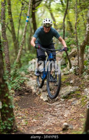Ciclista esperto percorre un sentiero boschivo roccioso su una mountain bike blu, indossa un casco, guanti e ginocchiere. Scatto d'azione che trasmette concentrazione, avventura Foto Stock