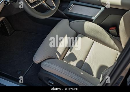 Colpo ad angolo alto di un sedile anteriore in pelle perforata beige chiaro, del bordo del volante e della console centrale di un'auto moderna parcheggiata all'aperto alla luce del giorno. Foto Stock