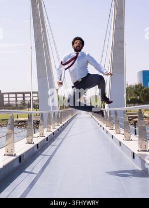 Uomo d'affari mediorientale in abito salta su un ponte moderno, celebrando il successo di una startup e un risultato aziendale sicuro di sé. Energetico, professionista sorridente Foto Stock