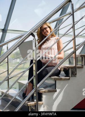 Giovane donna d'affari del Medio Oriente seduta sulle scale dell'ufficio che usa un laptop, sorride con sicurezza in un ambiente moderno di startup aziendale. Professore casual Foto Stock