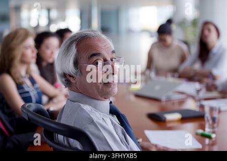 Un dirigente anziano con occhiali ascolta attentamente durante una riunione di lavoro in una sala conferenze moderna e luminosa, circondato da colleghi, laptop Foto Stock