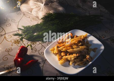 Una fotografia di natura morta ad angolazione alta posizionata su un tavolo da cucina coperto da una tovaglia a motivi. Un piatto quadrato bianco contiene una porzione di cibo appena cotto Foto Stock