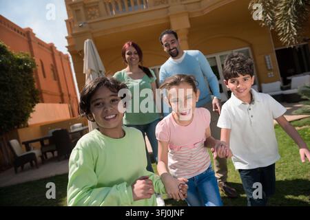 Una famiglia araba felice del Medio Oriente si diverte a divertirsi nel giardino, mentre i genitori sorridono mentre tre bambini corrono verso la macchina fotografica, mostrando gioia e divertimento Foto Stock