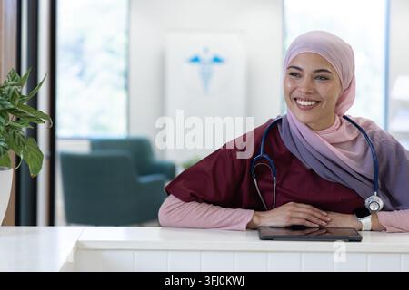 Infermiera donna in scrub di midollo seduto alla scrivania della clinica, sorridente, utilizzando una compressa con stetoscopio. Reception, hall, assistenza sanitaria, accogliente, luce naturale, ottanio, Foto Stock