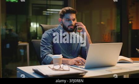 Uomo d'affari arabo concentrato e serio che parla al telefono, lavora con il laptop in ufficio la sera, uomo d'affari indù indiano sovraccarico, agente maschile parla Foto Stock