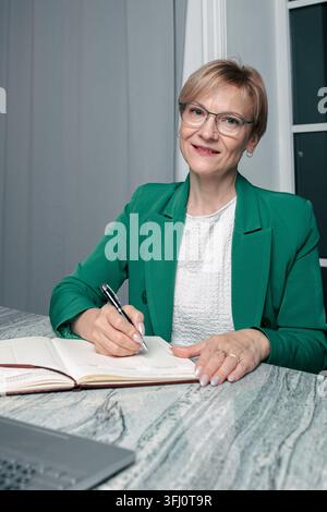 Una donna d'affari matura con un blazer verde siede alla scrivania scrive appunti in un planner guarda all'espressione che si adatta alla fotocamera sullo sfondo morbido dell'ufficio Foto Stock