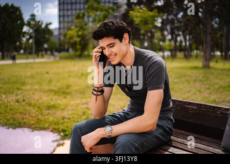 Giovane uomo che parla al telefono con lo smartphone mentre è seduto nel parco Foto Stock