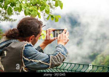 Uomo caucasico vista posteriore che scatta una foto con lo smartphone di un paesaggio montano nebbioso in giacca di jeans durante una vacanza all'aperto Foto Stock