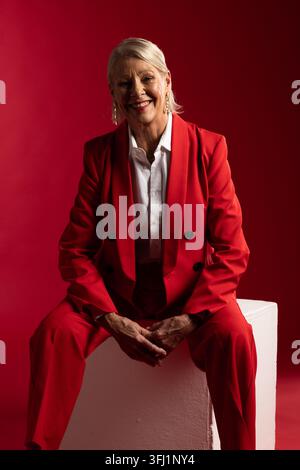 Donna anziana sorridente e seduta su un piedistallo bianco in studio, indossa un blazer e pantaloni rossi Foto Stock