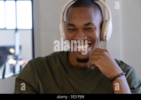 Uomo adulto afroamericano sorridente alla scrivania in ufficio con cuffie bianche e braccialetto di mento. Ritratto, cuffia, barba, verde oliva, coworking, Foto Stock