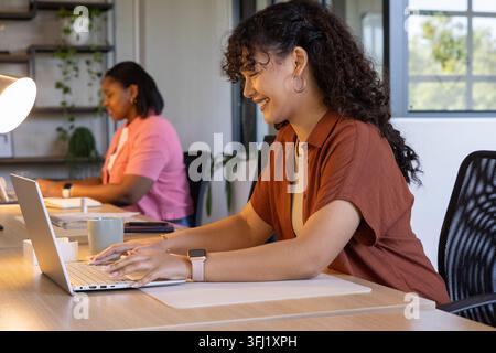 Colleghi diversi che lavorano in un ufficio moderno, donna ispanica che scrive su un laptop con una tazza verde nelle vicinanze. Coworking, collaborazione, scrivania, produttività, dayli Foto Stock