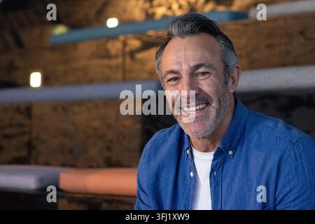 Uomo di mezza età sorridente, seduto sulla panchina cuscini nella lounge con camicia blu, spazio per le copie. Interni, caldi, ambiente, legno, rilassato, casual, maturo Foto Stock