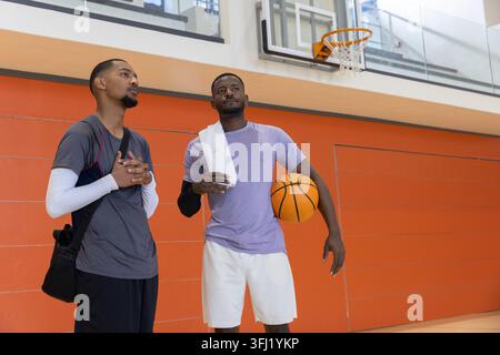 Diversi amici maschi che chiacchierano sul campo da basket in palestra con attrezzatura sportiva che tiene la palla vicino al cerchio. Atletica, cameratismo, conversazione, struttura, vibrante, o Foto Stock