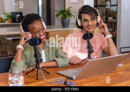 Diversi co-host seduti nella cucina di casa registrano con laptop, microfoni e cuffie, sorridendo. Podcast, duo, studio, collaborazione, conversazione Foto Stock