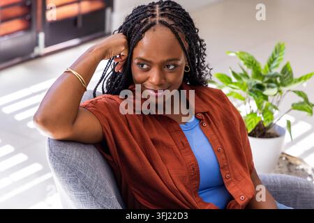 Donna africana seduta, camicia arrugginita con bracciale a cerchio vicino a una poltrona, doghe che proiettano luce sul pavimento di piastrelle. Sedia, casa, interno, vita Foto Stock