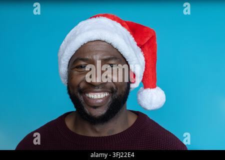 Uomo afro-americano che sorride in grande, indossa un cappello di Babbo Natale rosso e un maglione a maglia marrone in studio. Turchese, sfondo, barba, baffi, pelliccia, pompon, happ Foto Stock