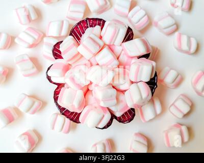 Vista dall'alto di mini marshmallow rosa e bianchi a strisce in una ciotola e sparsi su uno sfondo bianco. Foto Stock