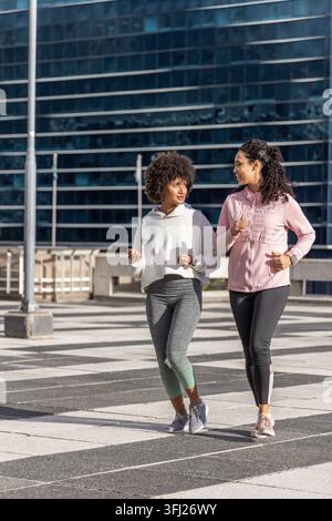 Diverse amiche che camminano attraverso la piazza a strisce, attraverso un edificio di vetro, indossando scarpe da running. Urban, outdoor, duo, sneakers, athleisure, lavoro Foto Stock