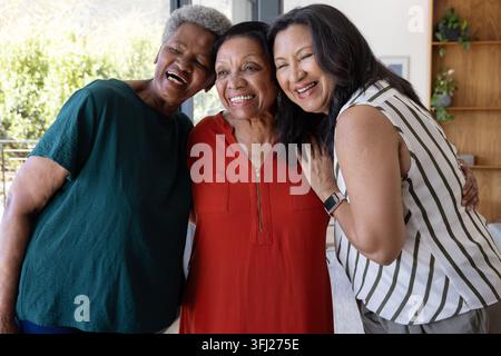 Diverse amiche anziane che abbracciano e ridono nel soggiorno con abito rosso e smartwatch. Trio, compagnia, calore, luce solare, interni, casa, Foto Stock
