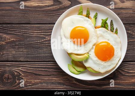 Colazione gustosa. Uova fritte con asparagi verdi e avocado su pane tostato sul piatto su un tavolo di legno. Vista dall'alto Foto Stock