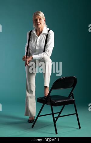 Donna anziana in posa in uno studio verde acqua con il piede su una sedia nera, indossa una camicia bianca e bretelle Foto Stock