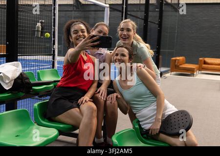 Diverse amiche in posa per un selfie al campo del padel con un abbigliamento sportivo che regge smartphone e racchetta. Compagni di squadra, gruppo, gioia, vibrante, colorato, social, Foto Stock