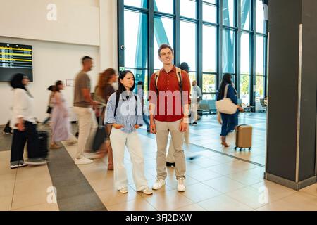 Coppia di giovani adulti in piedi in un terminal aeroportuale affollato mentre altri passeggeri passano Foto Stock