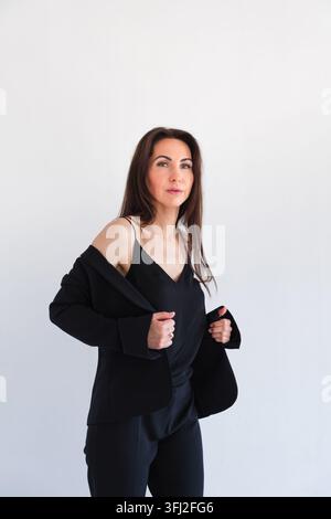 Donna caucasica in un completo da lavoro nero si trova contro un muro bianco, guardando la fotocamera con un'espressione sicura di sé. Abbigliamento professionale, business Foto Stock