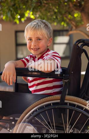 Ragazzo seduto su una sedia a rotelle nel cortile, indossa una maglietta a righe rosse e bianche, sorride. All'aperto, gioventù, mobilità, accessibilità, s Foto Stock