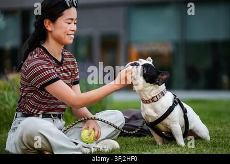 Giovane donna adulta che regala una deliziosa delizia al suo bulldog francese in un parco Foto Stock
