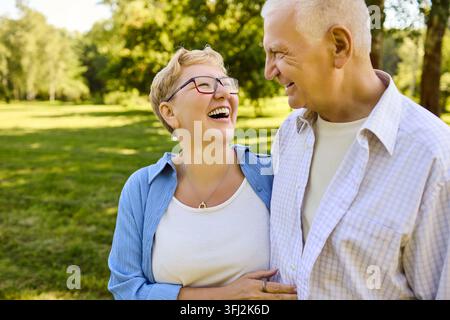 Coppia di anziani felici che ridono e si abbracciano in un parco soleggiato, amore in pensione Foto Stock