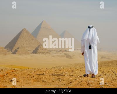 Un uomo arabo in un thobe bianco e keffiyeh si trova in piedi, osservando le antiche piramidi di Giza attraverso il deserto. Una luce diurna serena e brumosa evoca meraviglia ed eredità Foto Stock