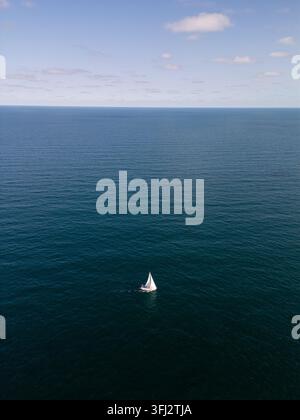 Vista drone di una solitaria barca a vela bianca che naviga attraverso un mare calmo e blu profondo sotto un cielo sereno. Ampia paesaggio aereo panoramico di una nave a vela Foto Stock