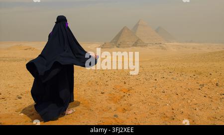 Una donna araba velata in un'abaya nera che scorre guarda verso le piramidi di Giza attraverso il nebbioso deserto egiziano. Ambientazione tranquilla e senza tempo, ideale per tou egiziani Foto Stock
