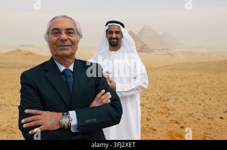 Un uomo d'affari arabo anziano con un vestito e un giovane emirato sorridente in kandura bianca si trovano davanti alle piramidi di Giza in Egitto. Autentica MENA Foto Stock