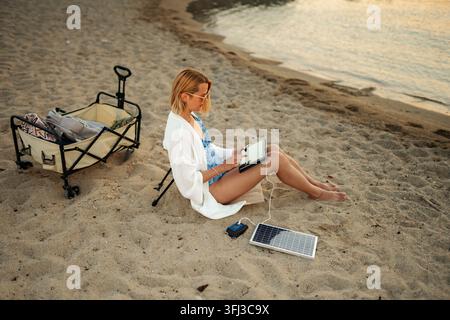 Donna che utilizza un tablet alimentato da un pannello solare portatile mentre si rilassa sulla sabbia Foto Stock