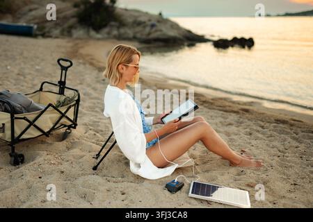 Donna che si gode il tramonto sulla spiaggia, ricaricando il suo tablet con un pannello solare portatile Foto Stock