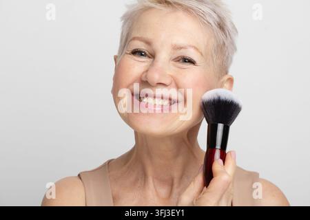 Ritratto di una bella donna anziana sorridente che si trucca, applica il rossore sulla guancia e guarda verso la fotocamera Foto Stock