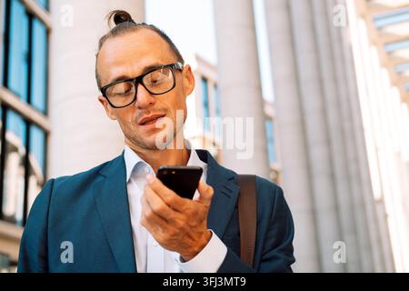 Giovane uomo affascinante con occhiali che registra un messaggio audio sullo smartphone mentre cammina per la strada della città. Foto Stock