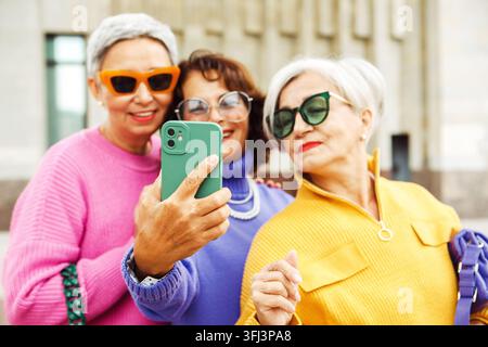 Tre donne anziane che si scattano un selfie insieme all'aperto, si divertono e sorridono indossando abiti colorati e occhiali da sole, fotografia ad alta risoluzione Foto Stock