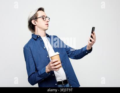 Giovane uomo che scatta un selfie e tiene una tazza di caffè da asporto su sfondo bianco Foto Stock