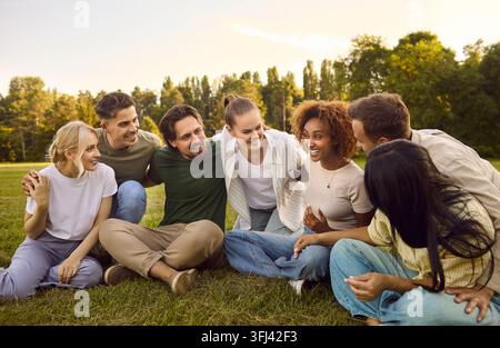 Amici diversi e felici che chiacchierano e ridono seduti insieme sull'erba nel parco Foto Stock