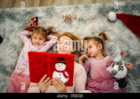 Madre che legge una storia di Natale a bambini felici su un tappeto soffice Foto Stock