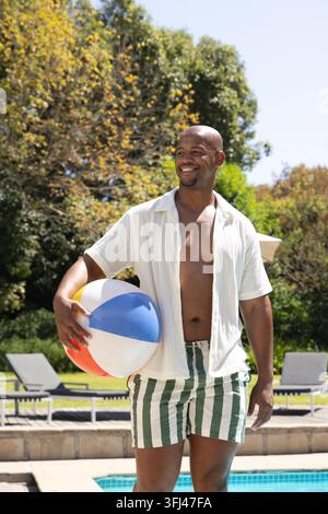 Uomo afroamericano in posa in piscina con un pallone da spiaggia colorato, indossa una camicia e un costume a righe. Pallone da spiaggia, soleggiato, all'aperto, bordo piscina, tempo libero, vacanza Foto Stock