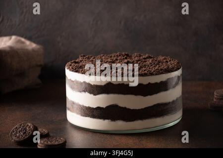 Torta trifle fatta in casa a strati con briciole di biscotti al cioccolato e panna prima della decorazione di Halloween, circondata da biscotti su un tavolo rustico scuro. Foto Stock