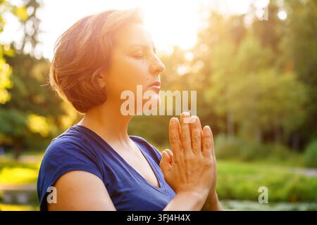 Primo piano di una donna caucasica matura che prega con le mani giunte nella calda luce della sera, ideale per contenuti su fede, gratitudine e consapevolezza. Foto Stock