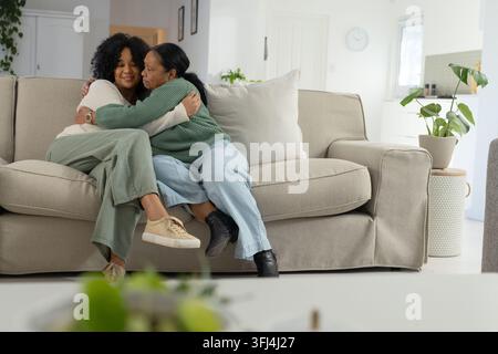 Madre e figlia afroamericane anziane sedute su un divano beige che si abbracciano con una pianta nel soggiorno Foto Stock