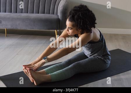 Donna che si allunga in avanti su un tappeto da yoga scuro nel soggiorno, indossa una canottiera grigia e uno smartwatch. Fitness, casa, benessere, flessibilità Foto Stock