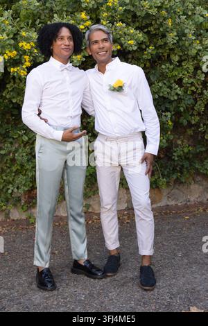Partner maschi afroamericani in piedi e sorridenti vicino a una siepe, con camicie bianche e fiore all'occhiello. Coppia, romanticismo, matrimonio, giardino, fiori, petali Foto Stock